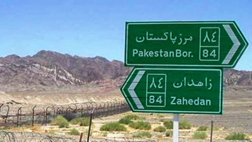 A long, high, barbed-wire fence near the border between Iran and Pakistan. Behind the fence is a range of low mountains. In front of the fence, in the foreground, are two green road signs, with text in Persian and Roman letters. The lower sign, pointing left, reads "Zahedan". The upper sign, pointing right, reads "Pakestan Bor.", short for "Border". The Persian text begins with the word "marz", in Arabic letters, which is Persian for "border".