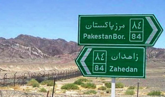 A long, high, barbed-wire fence near the border between Iran and Pakistan. Behind the fence is a range of low mountains. In front of the fence, in the foreground, are two green road signs, with text in Persian and Roman letters. The lower sign, pointing left, reads "Zahedan". The upper sign, pointing right, reads "Pakestan Bor.", short for "Border". The Persian text begins with the word "marz", in Arabic letters, which is Persian for "border".